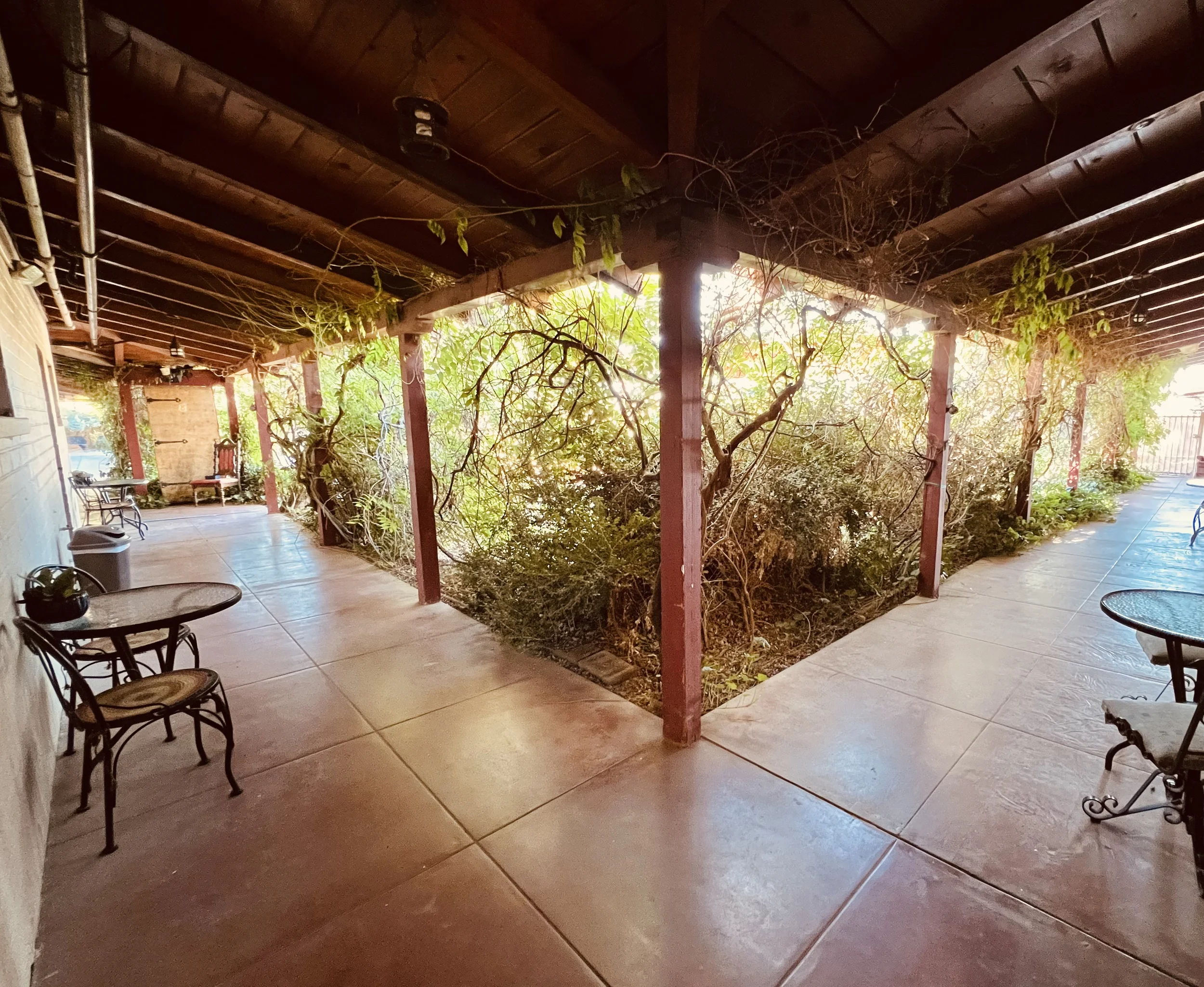 the corner of  the walk way with vines on one side and hotel room doors and table and chairs on the other. 